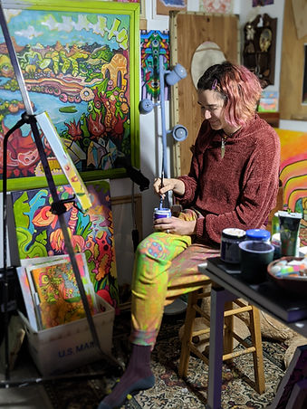 Rae Grand in her studio next to an easle with colorful work behind her
