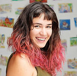 portrait of the artist, smiling with long poofy brown hair and short bangs watercolors hang behind her