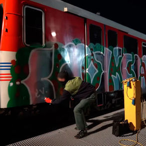 Graffiti en cours de réalisation sur un train dans un dépôt ferroviaire, avec un artiste peignant une grande pièce colorée à la bombe de peinture spray sur un wagon.