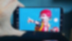 Hand holding a smartphone displaying a toy figure in a red cap pointing against a blue background. Camera app interface is visible.
