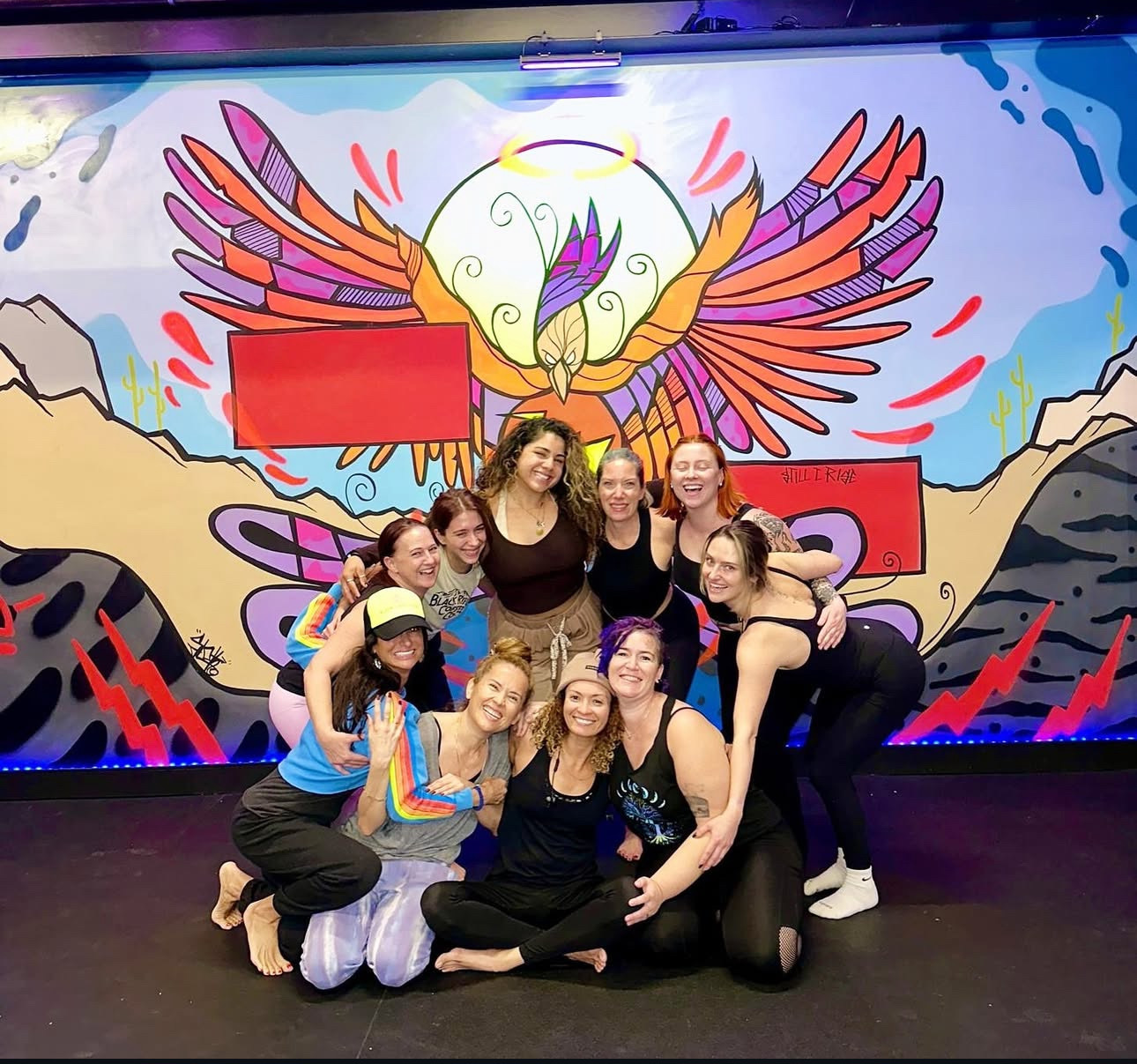 Smiling yogis pose before colorful wing mural after class at Black Swan Yoga Midtown Phoenix.