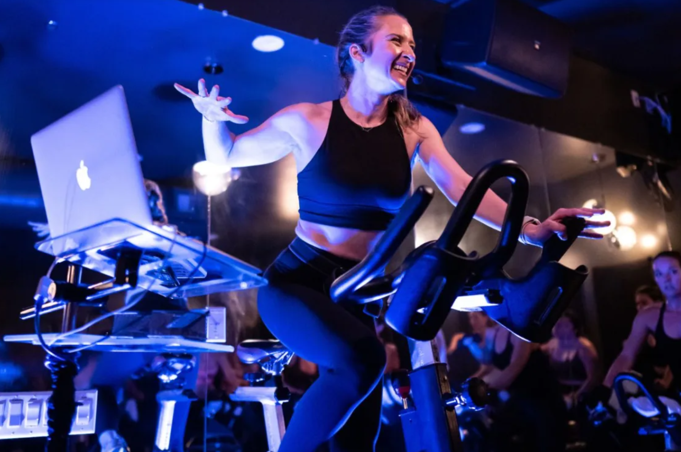 Indoor cycling instructor at Motiv Fitness Austin riding a bike on stage next to a DJ booth under blue lights.