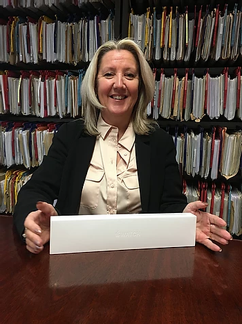 A smiling person sits at a table with an Apple Watch. Background shows shelves of organised coloured folders.