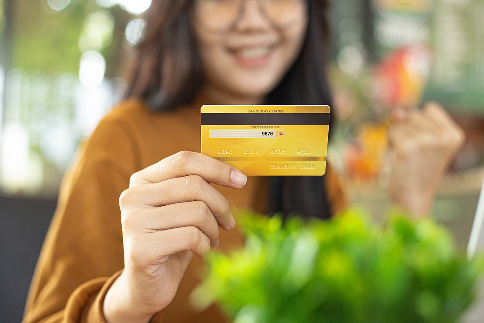A cheerful Asian woman holding a yellow credit card and smiling while sitting at a table in a cozy indoor environment. She gestures confidently with a fist pump, expressing success and satisfaction.