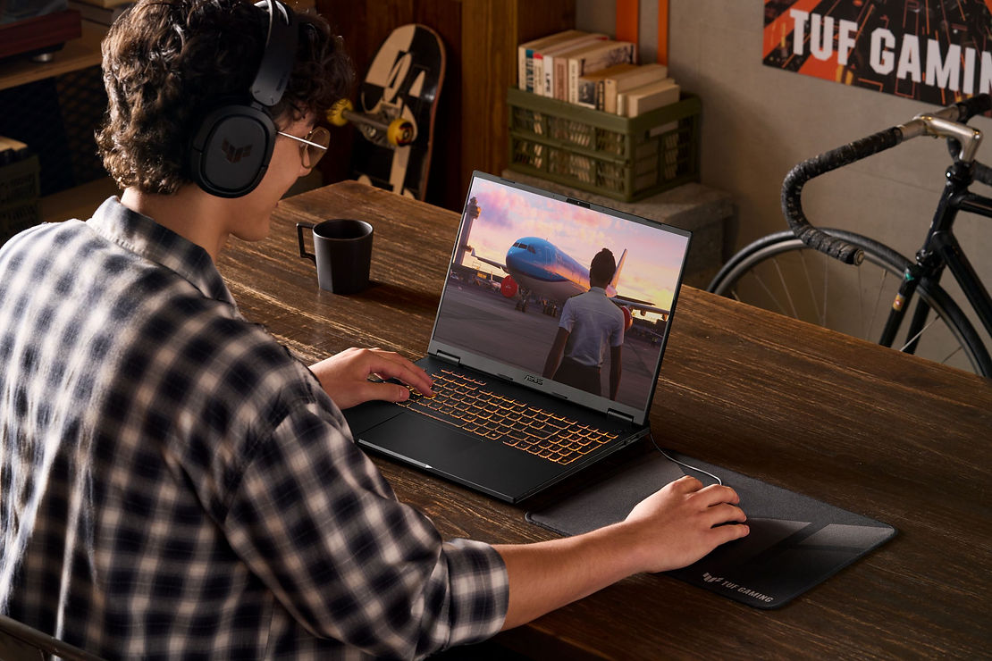 The image shows a person wearing a black shirt, sitting at a wooden desk, using an Asus TUF Gaming laptop with a vibrant outdoor scene displayed on the screen, and a bicycle leaning against the wall.AI-generated content may be incorrect.