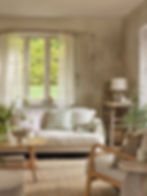 Cozy living room with a beige sofa, green and pink pillows, wooden table, lamp, and plants. Books titled "Home Comforts" on table. Airy vibe.