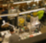 The photograph shows the SOS Eyewear Services workshop in Auckland. The photo shows two optical repair technicians expertly fixing glasses and sunglasses.