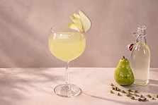 Pear & Cardamom Sparkler in a balloon glass next to a pear, clear swing-top bottle, and surrounded by scattered green cardamom pods against a pink backdrop.