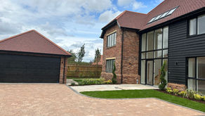 Modern two-story brick and black timber-clad house with large glass windows, red tiled roof, detached double garage, and landscaped driveway with fresh lawn and plants under a partly cloudy sky