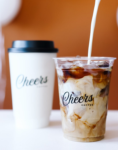 A hot Cheers Coffee cup next to an iced Cheers Coffee cup that's getting cream poured into it, creating a swirl effect