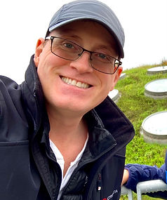 Mike similing in a grey baseball cap and black jacket in front of a green roof