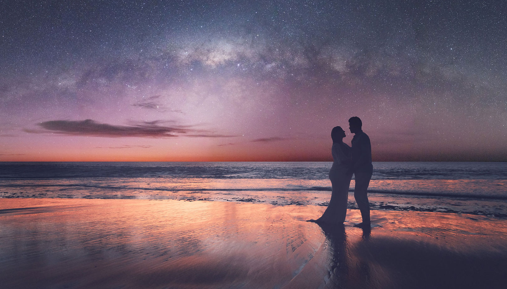 A couple, a man and pregnant women standing on the beach, they are silhouetted against the night sky with the bright milky way core behind and the last glimpses of sunlight reflecting on the wet sand