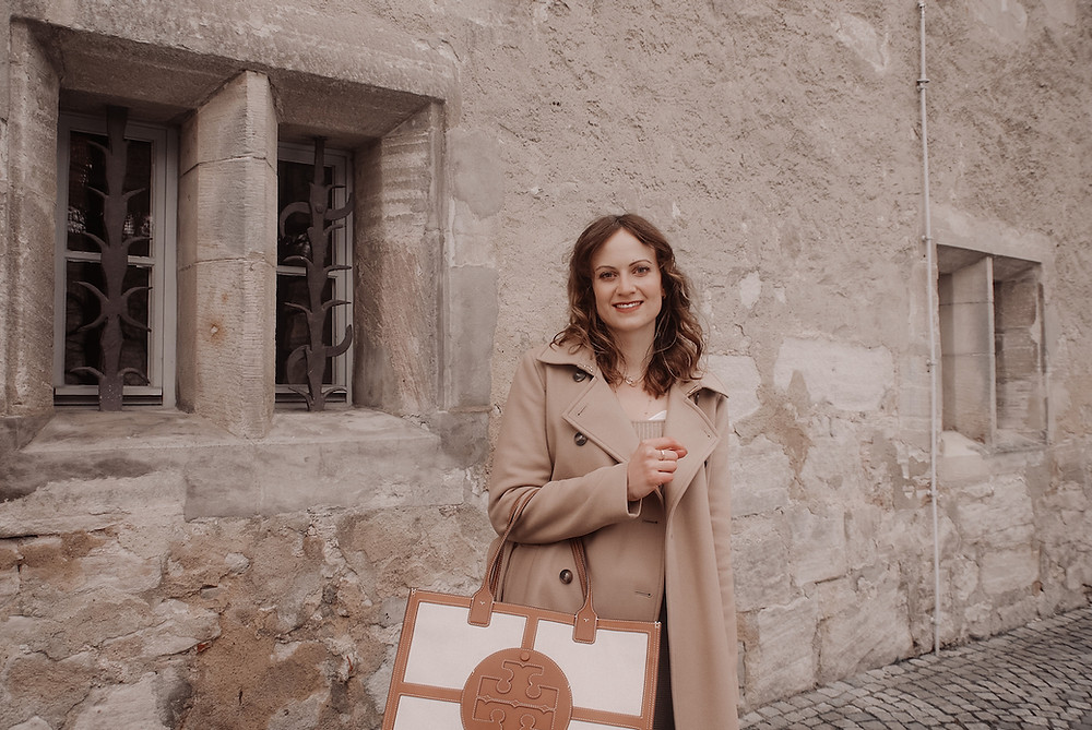 Junge Frau in einem beigen Mantel, mit großer Canvas Tasche und braunen Stiefeln vor einer Sandsteinwand