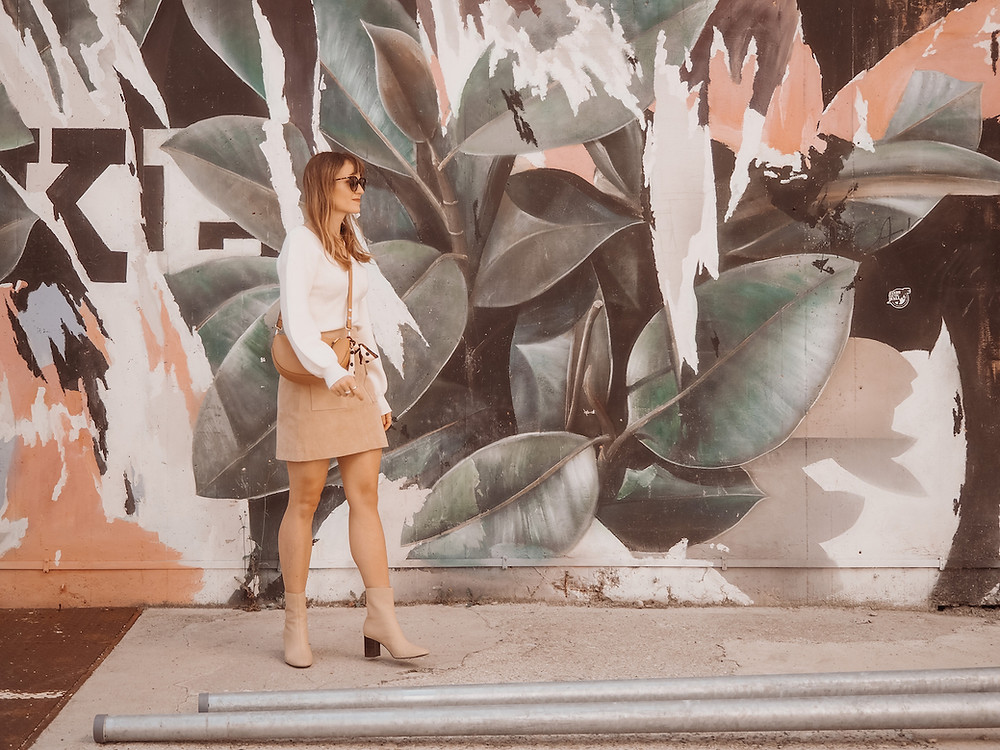 Young woman dressed in a trench coat, suede skirt, and white knit in front of a graffiti wall
