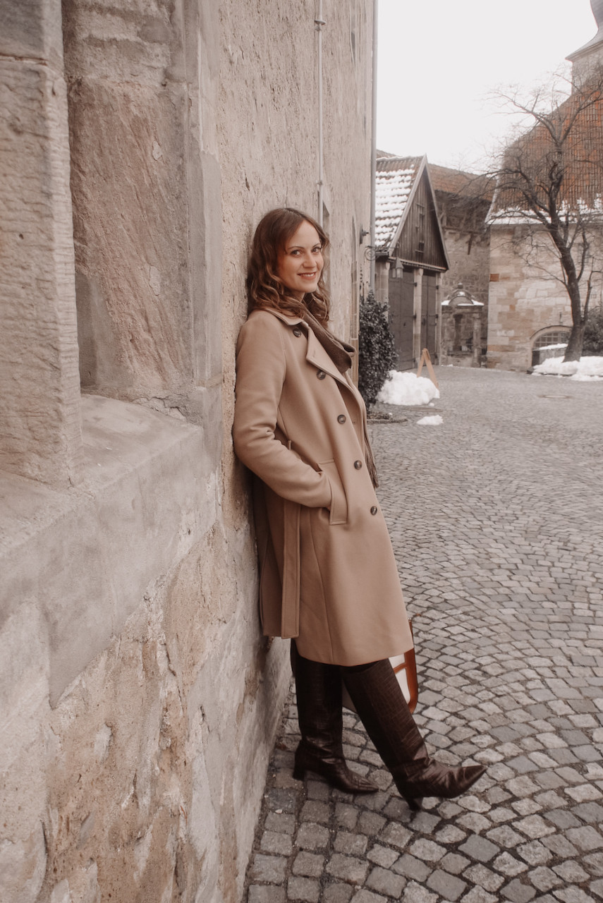 Junge Frau in einem beigen Mantel, mit großer Canvas Tasche und braunen Stiefeln vor einer Sandsteinwand
