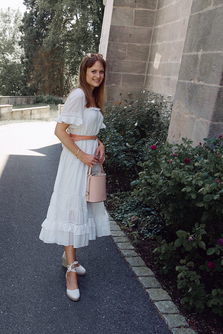 Young woman dressed in a midi length white summer dress, waist belt, white wedges, and pink bucket bag in front of a stone building