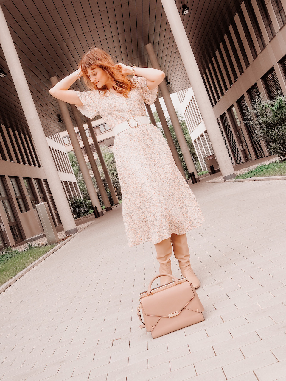 Young woman dressed in a floral midi dress, broad waist belt, beige boots and a brown bag in front of an office building