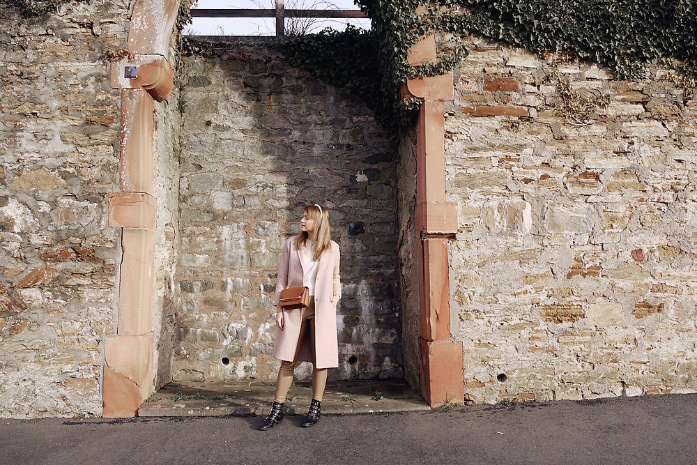 Girl in cream knit, tan colored faux leather pants, black boots, tan cross body bag, camel coat, and pearl head band standing in front of a wall