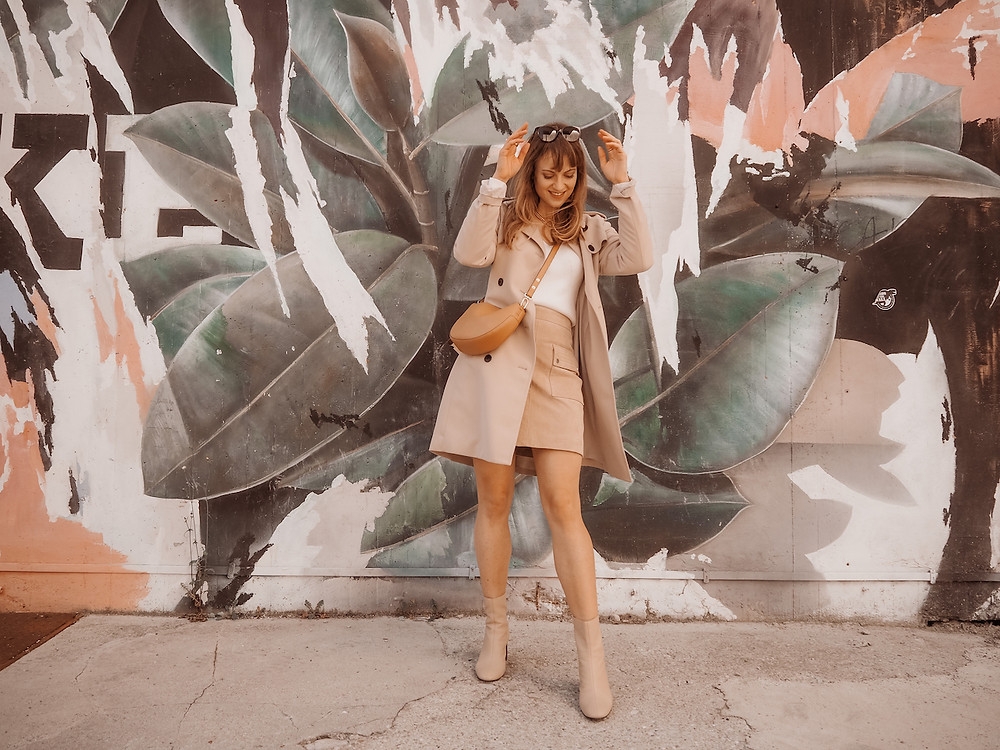Young woman dressed in a trench coat, suede skirt, and white knit in front of a graffiti wall