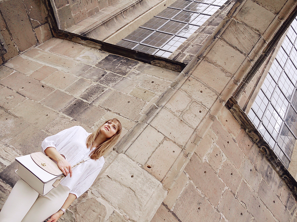 Young women dressed in white blouse, cream colored trousers, and white mules carrying a Coach bag, standing under a stone archway