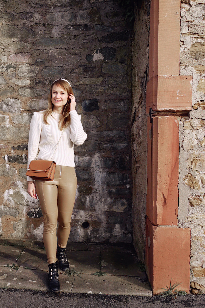 Girl in cream knit, tan colored faux leather pants, black boots, tan cross body bag, and pearl head band standing in front of a wall