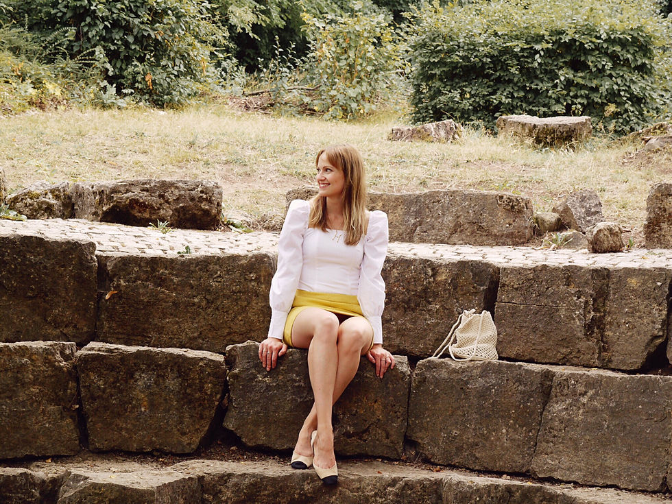 Girl in yellow skirt, white blouse and neutral accessories standing in front of a pile of stones