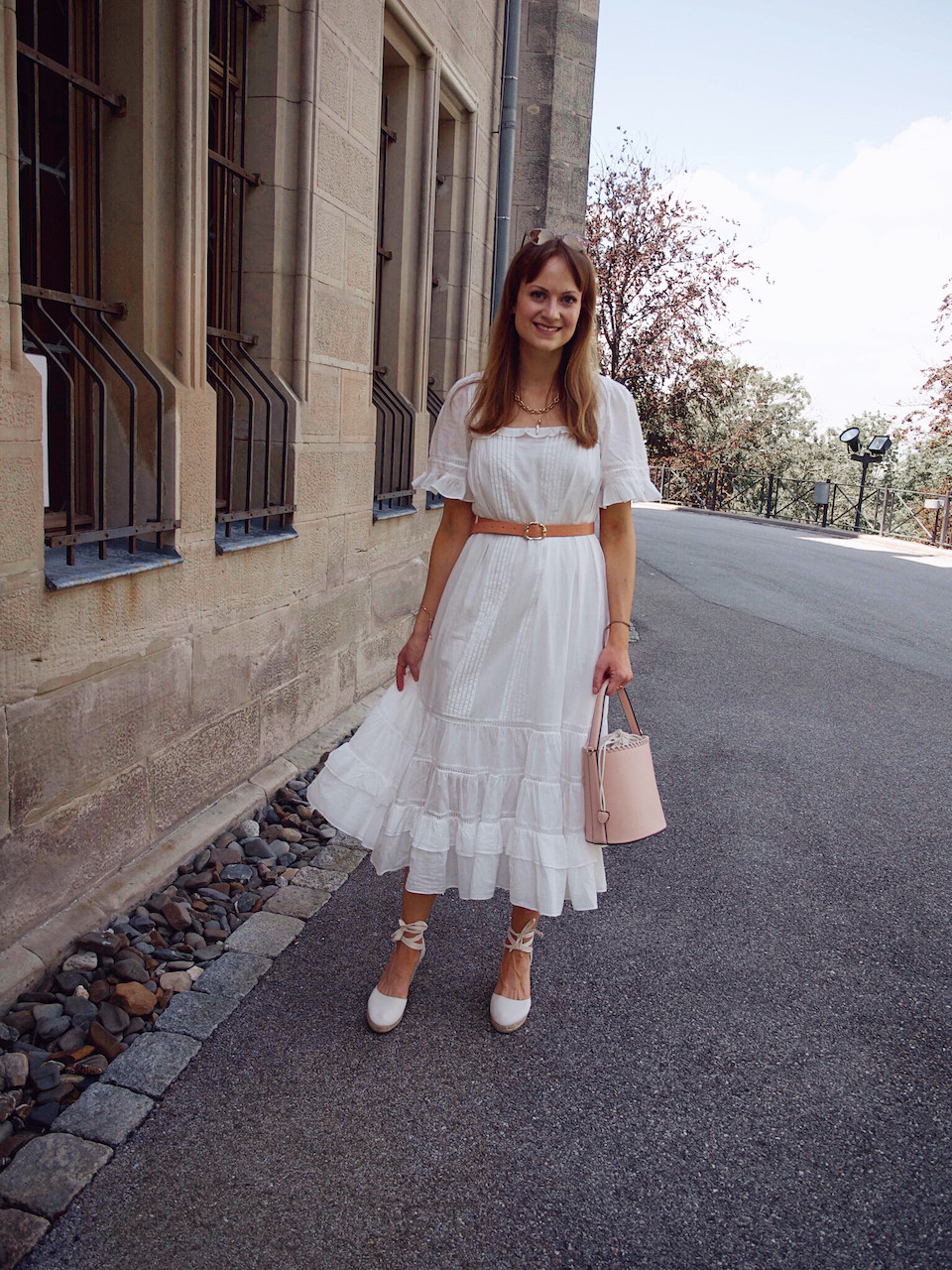Junge Frau in einem weißen Sommerkleid in Midilänge, weißen Keilabsatzschuhen und rosa Beuteltasche vor einem Steingebäude