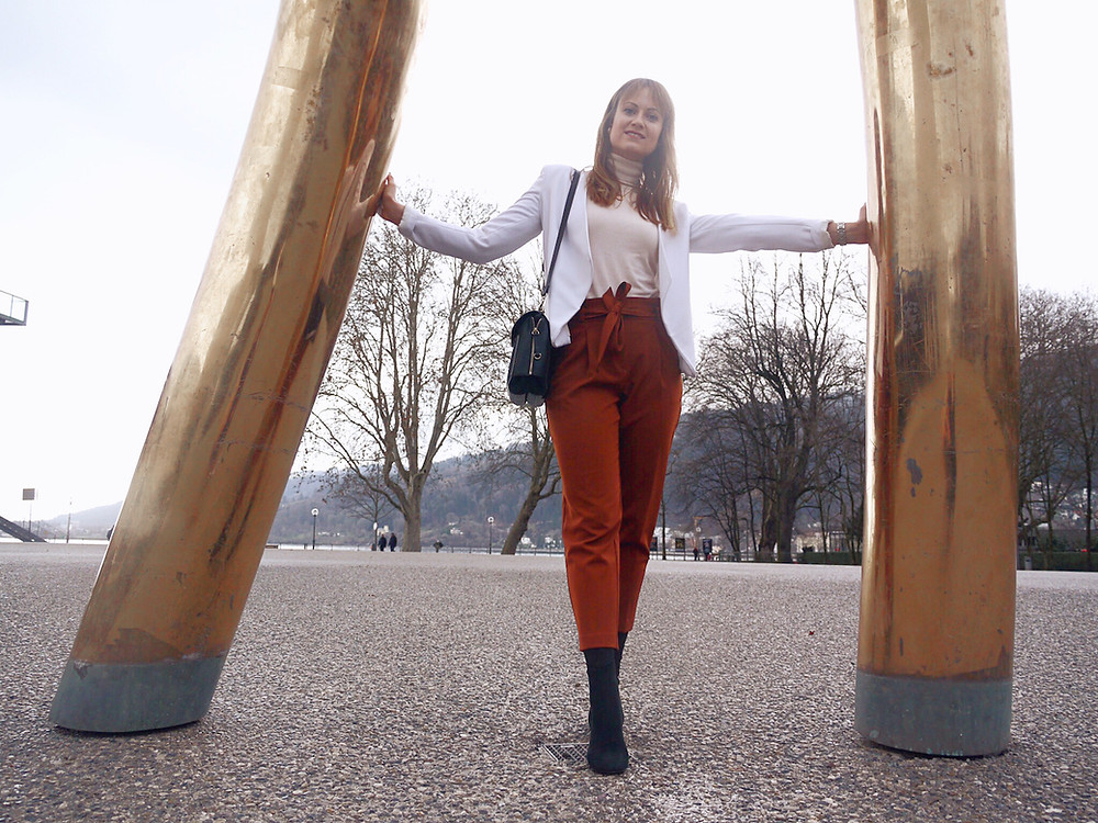 Girl in office outfit consisting of paperbag pants, roll neck jumper and white blazer standing between golden bars