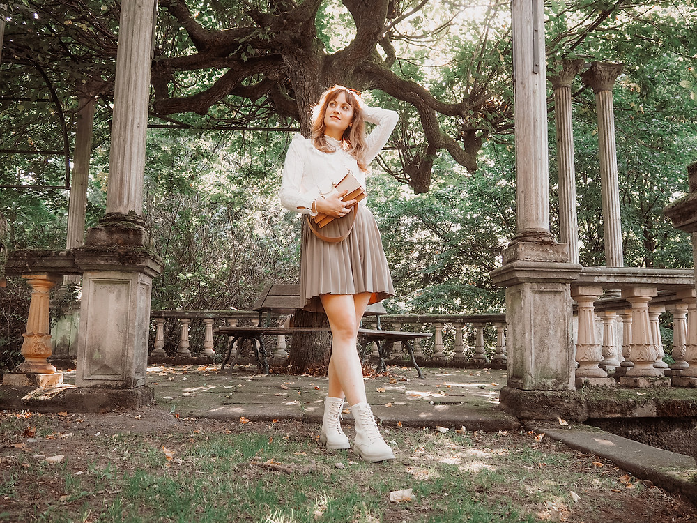 Young woman wearing a pleated mini skirt, trench coat, white lace blouse, white lace-up boots and Strathberry bag in a park