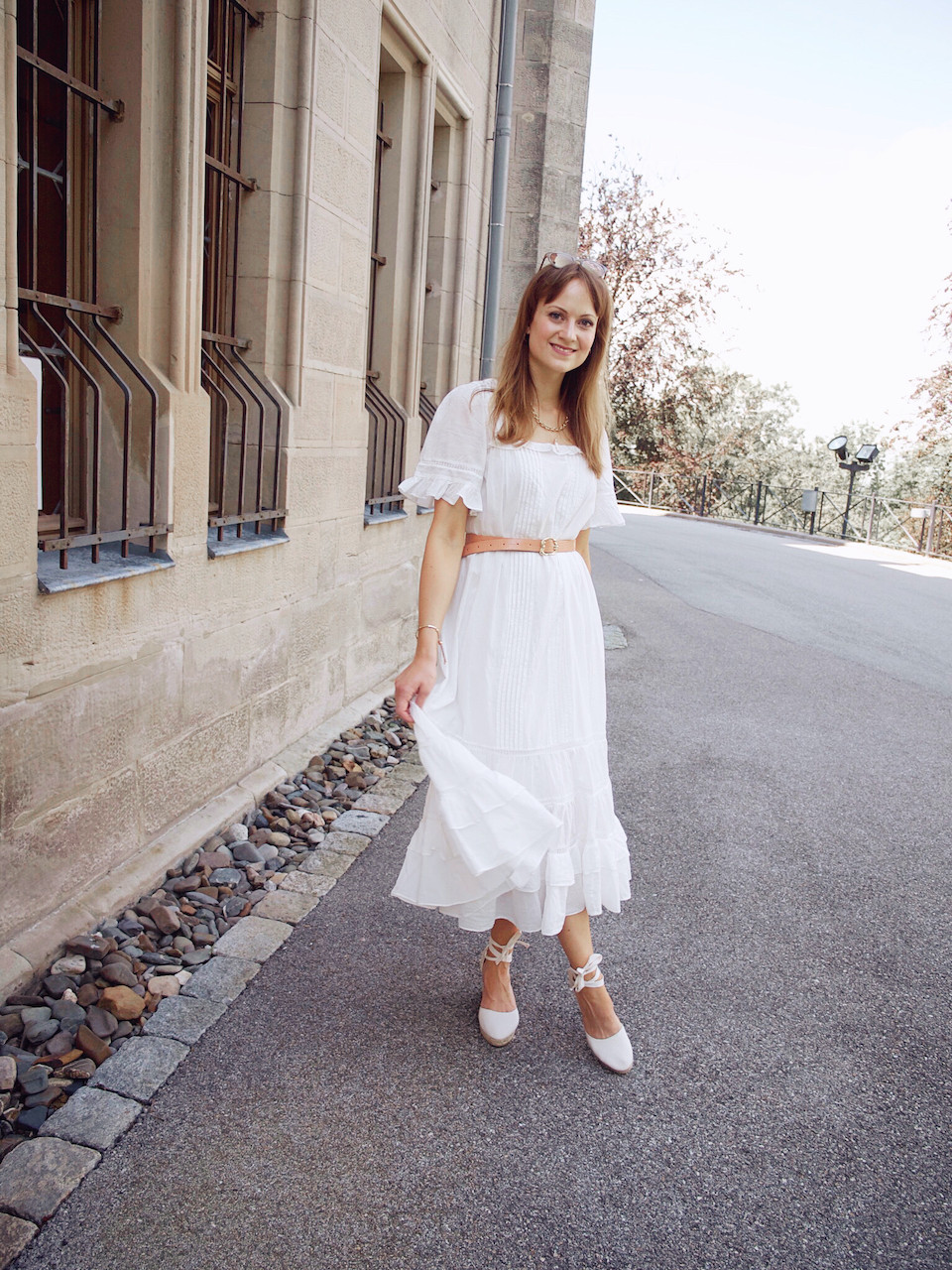 Young woman dressed in a midi length white summer dress, waist belt, white wedges, and pink bucket bag in front of a stone building