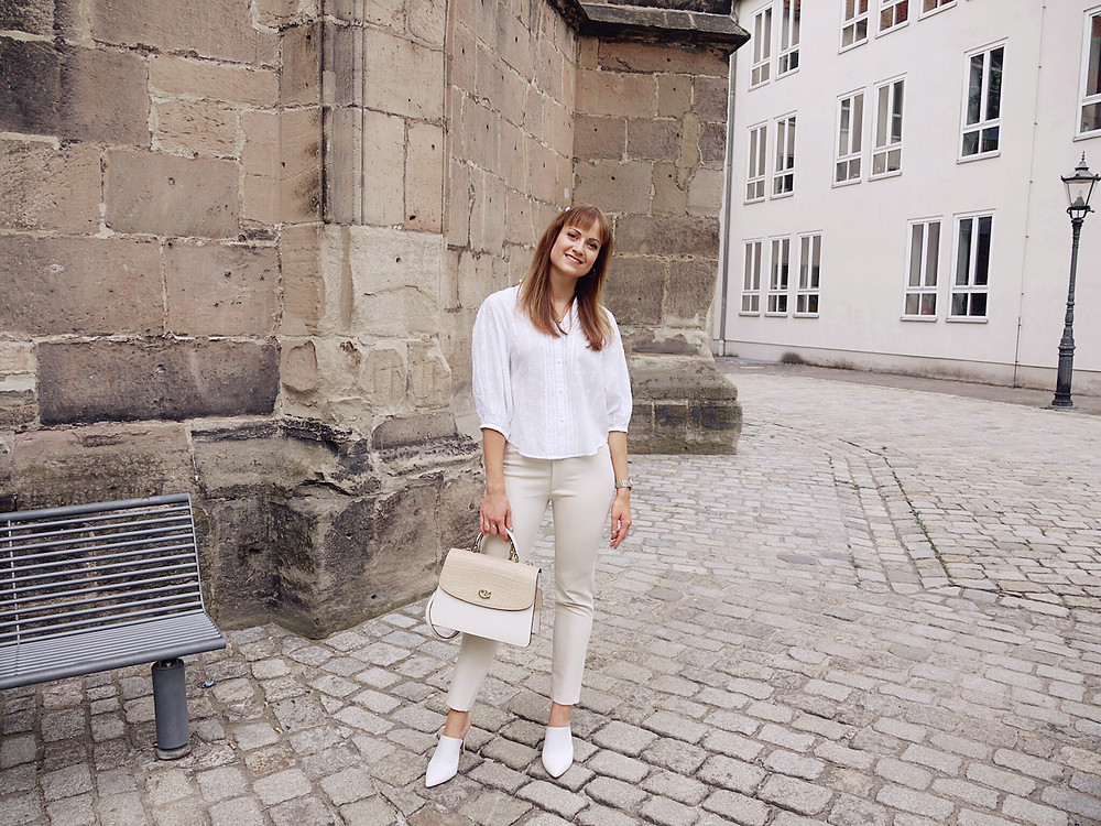 Junge Frau in einem Outfit aus weißer Bluse, cremefarbener Hose und weißen, spitzen Schuhen unter einem Steinbogen