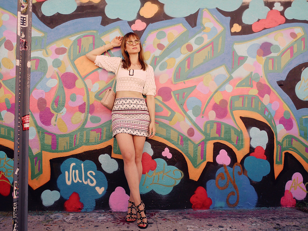 Girl in white lace crop top, patterned skirt, and studded sandals standing in front of a graffitti in Wynwood, Miami