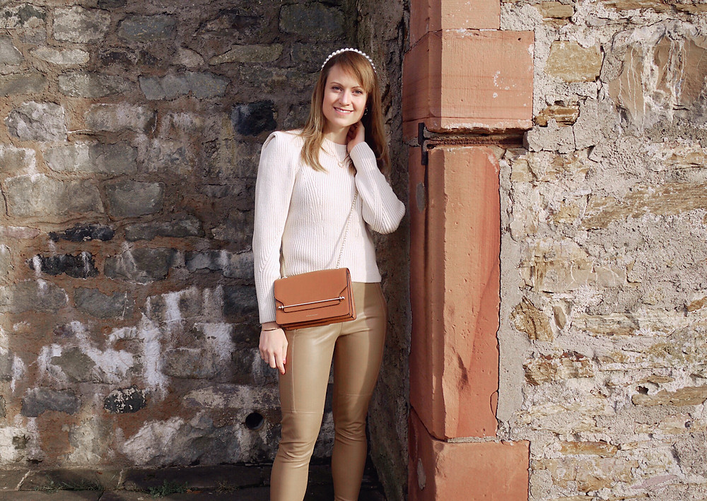 Girl in cream knit, tan colored faux leather pants, black boots, tan cross body bag, and pearl head band standing in front of a wall