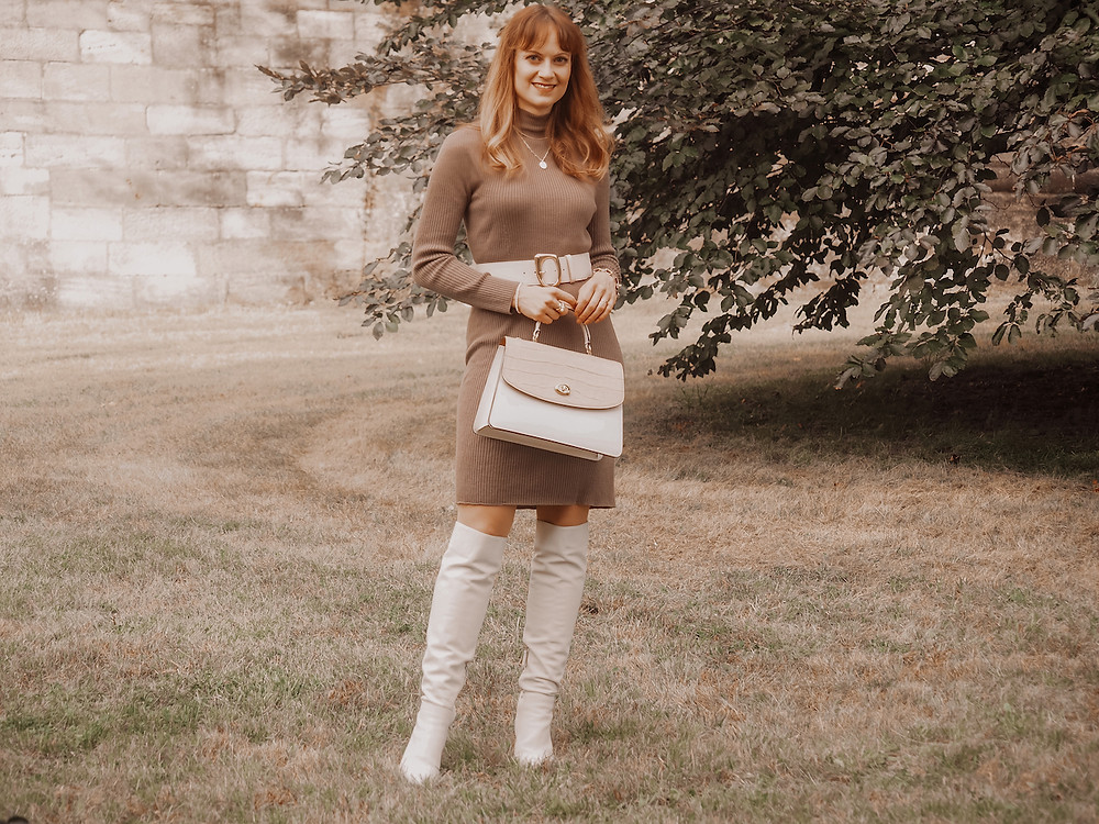 Young woman in a brown jumper dress, with white boots, a beige and white bag und white waist belt
