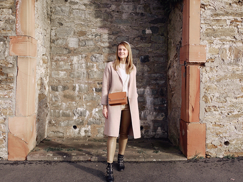 Girl in cream knit, tan colored faux leather pants, black boots, tan cross body bag, camel coat, and pearl head band standing in front of a wall