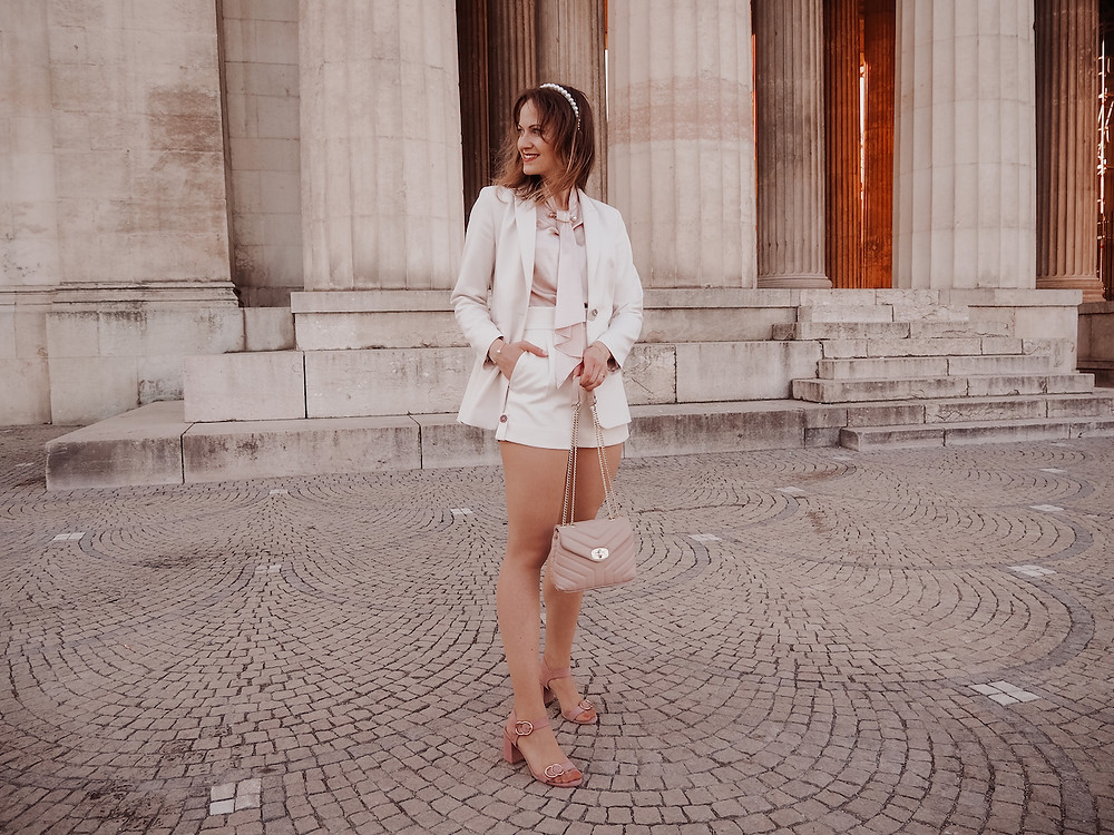 Young woman dressed in an outfit of a blazer, shorts, a silk blouse, and strappy sandals, standing in front of columns