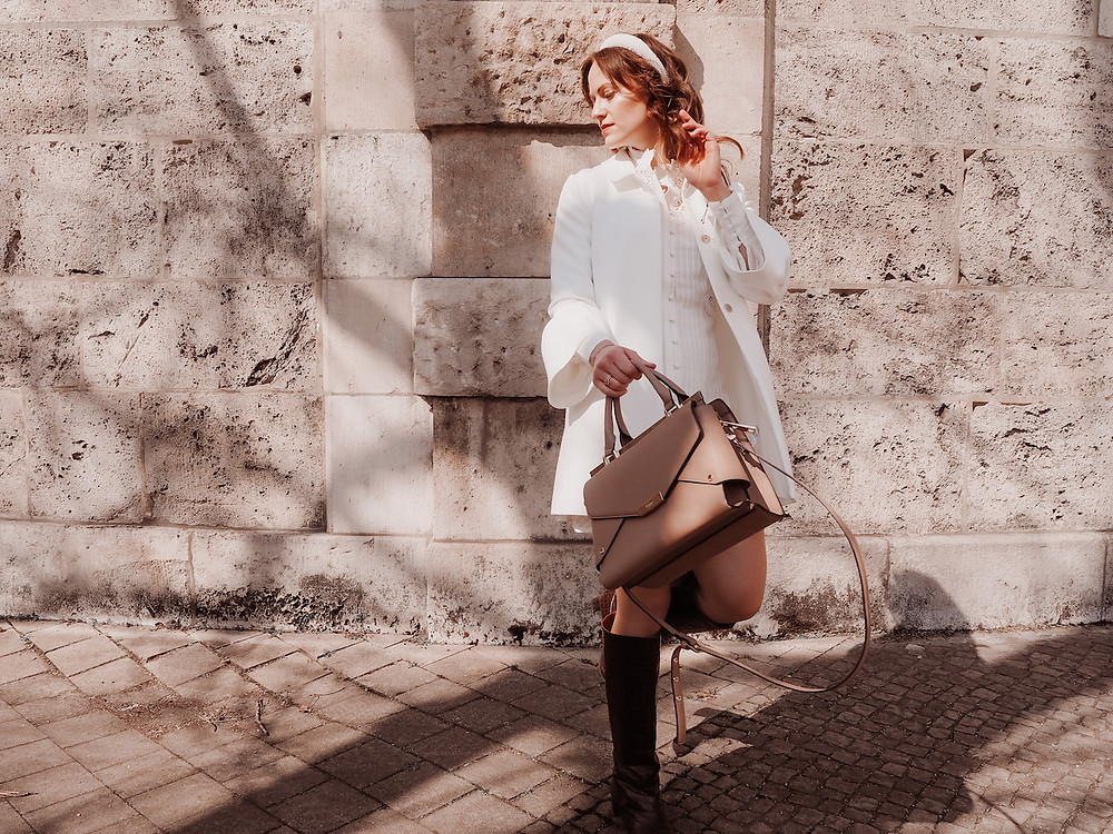 Junge Frau in einem Outfit aus weißem Spitzenkleid, weißem Mantel, braunen Stiefeln und einer braunen Tasche vor einer Steinwand