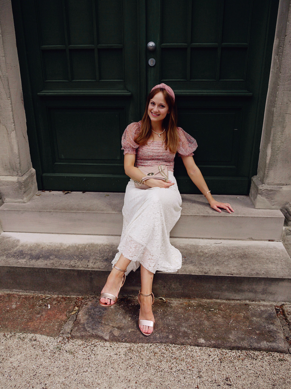 Young woman dressed in cream lace skirt, pink floral top and rose gold sandals in front of a dark green door