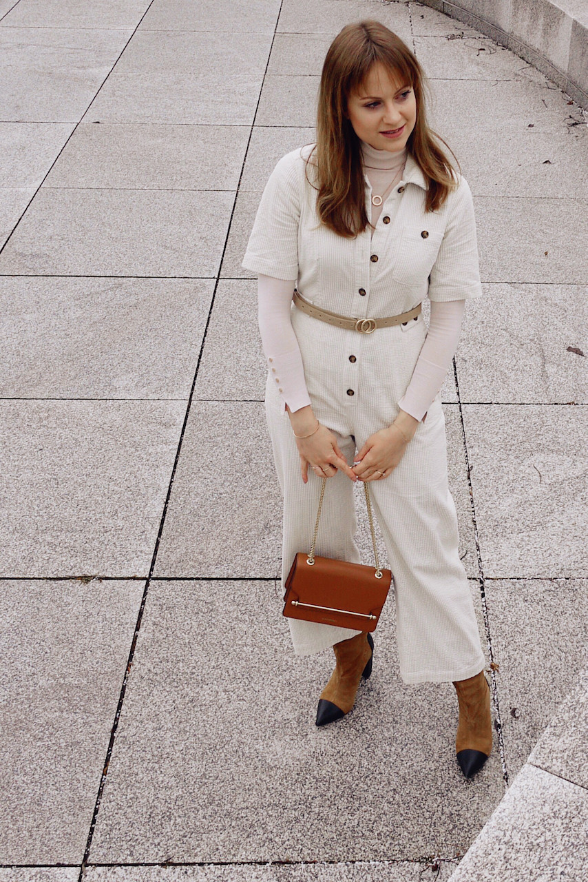 Young woman dressed in white jumpsuit, beige turtle neck and brown boots in front of stone steps