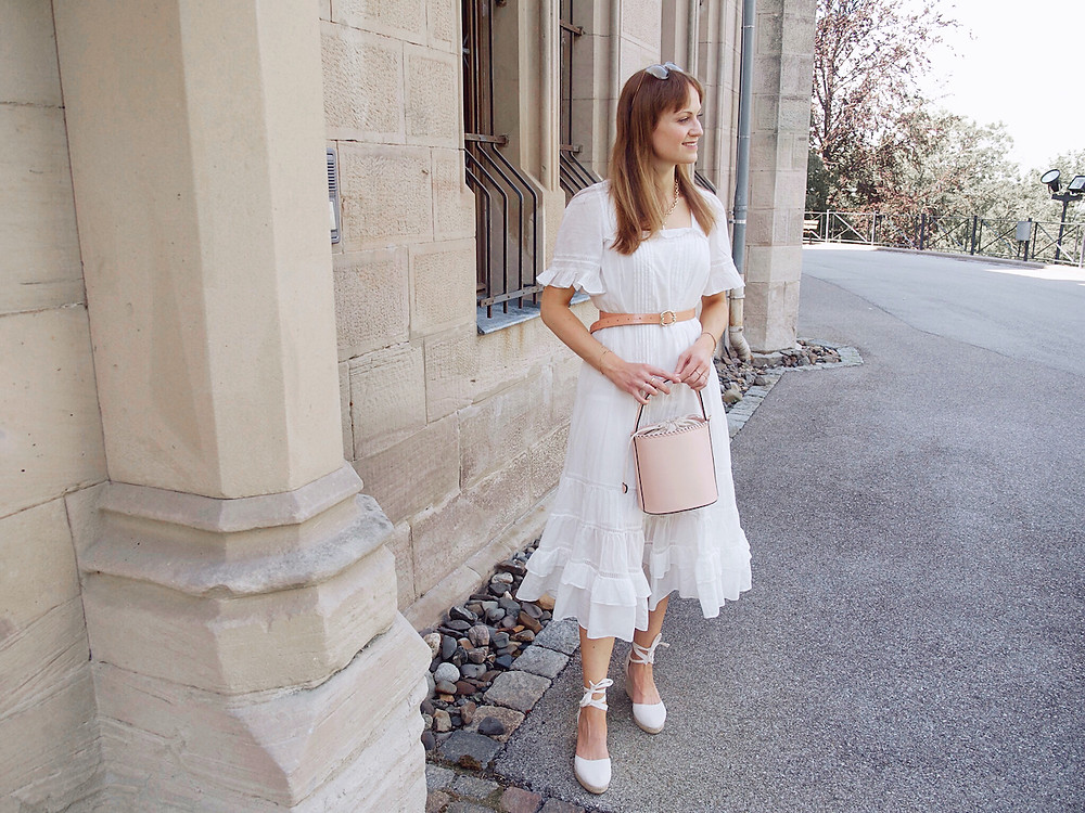 Junge Frau in einem weißen Sommerkleid in Midilänge, weißen Keilabsatzschuhen und rosa Beuteltasche vor einem Steingebäude