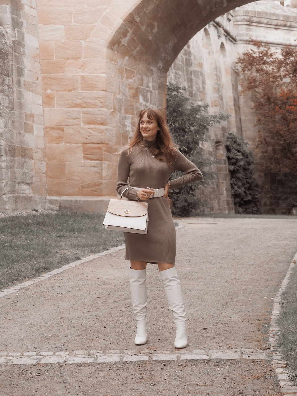 Young woman in a brown jumper dress, with white boots, a beige and white bag und white waist belt
