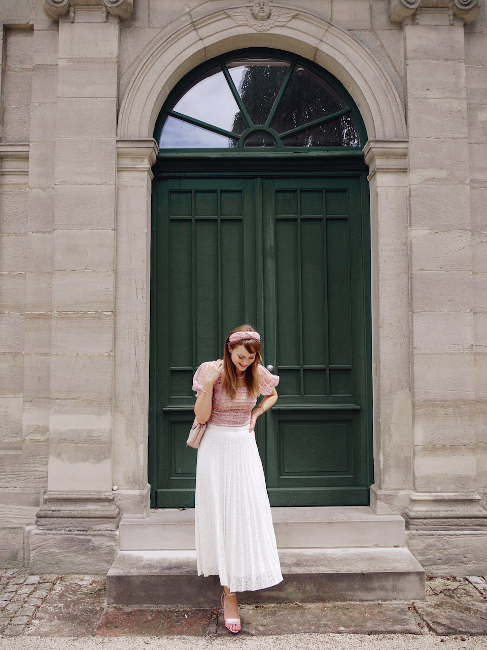 Young woman dressed in cream lace skirt, pink floral top and rose gold sandals in front of a dark green door