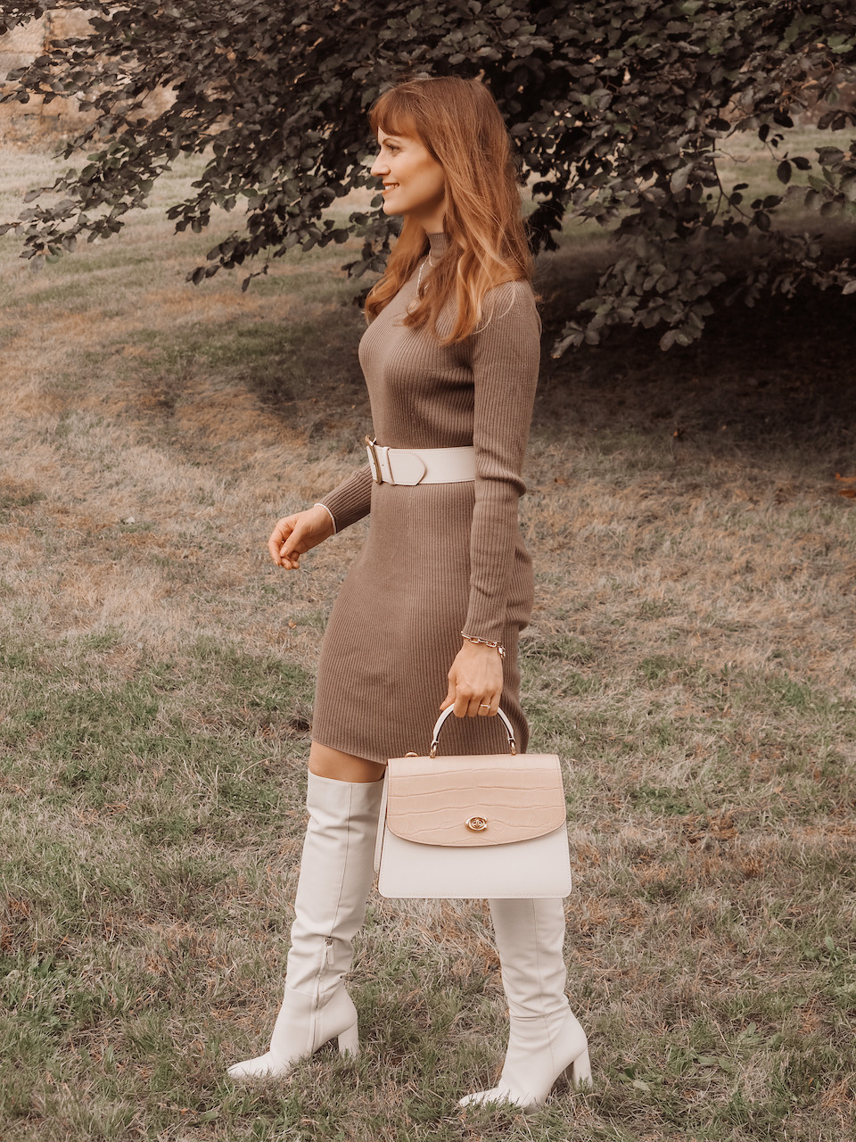 Junge Frau in einem braunen Pulloverkleid, weißen Stiefeln, weißem Taillengürtel und weiß-beider Coach Tasche vor einer Mauer mit Baum