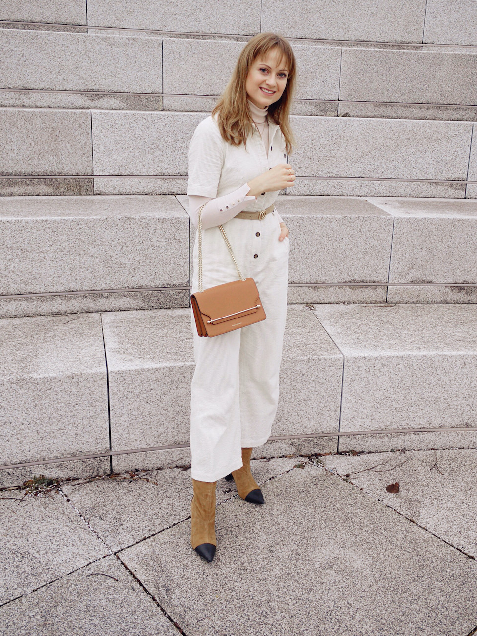 Young woman dressed in white jumpsuit, beige turtle neck and brown boots in front of stone steps