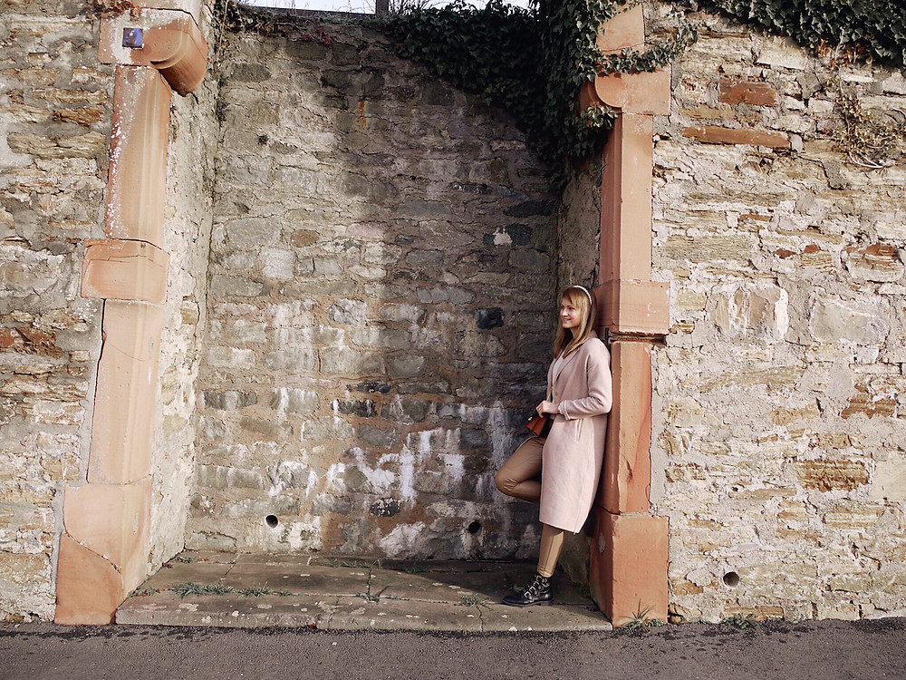 Girl in cream knit, tan colored faux leather pants, black boots, tan cross body bag, camel coat, and pearl head band standing in front of a wall