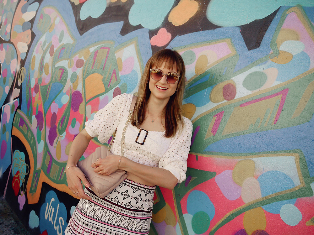 Girl in white lace crop top, patterned skirt, and studded sandals standing in front of a graffitti in Wynwood, Miami