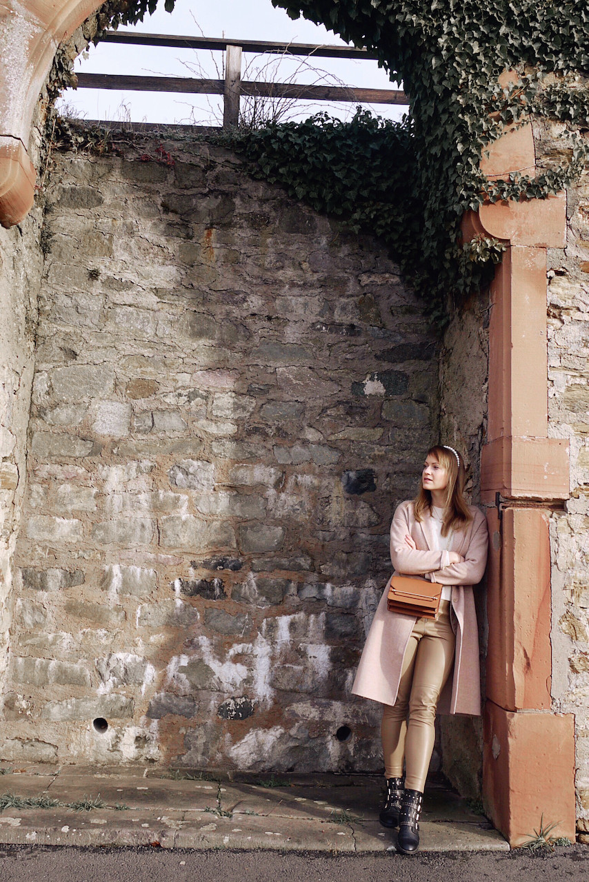 Girl in cream knit, tan colored faux leather pants, black boots, tan cross body bag, camel coat, and pearl head band standing in front of a wall