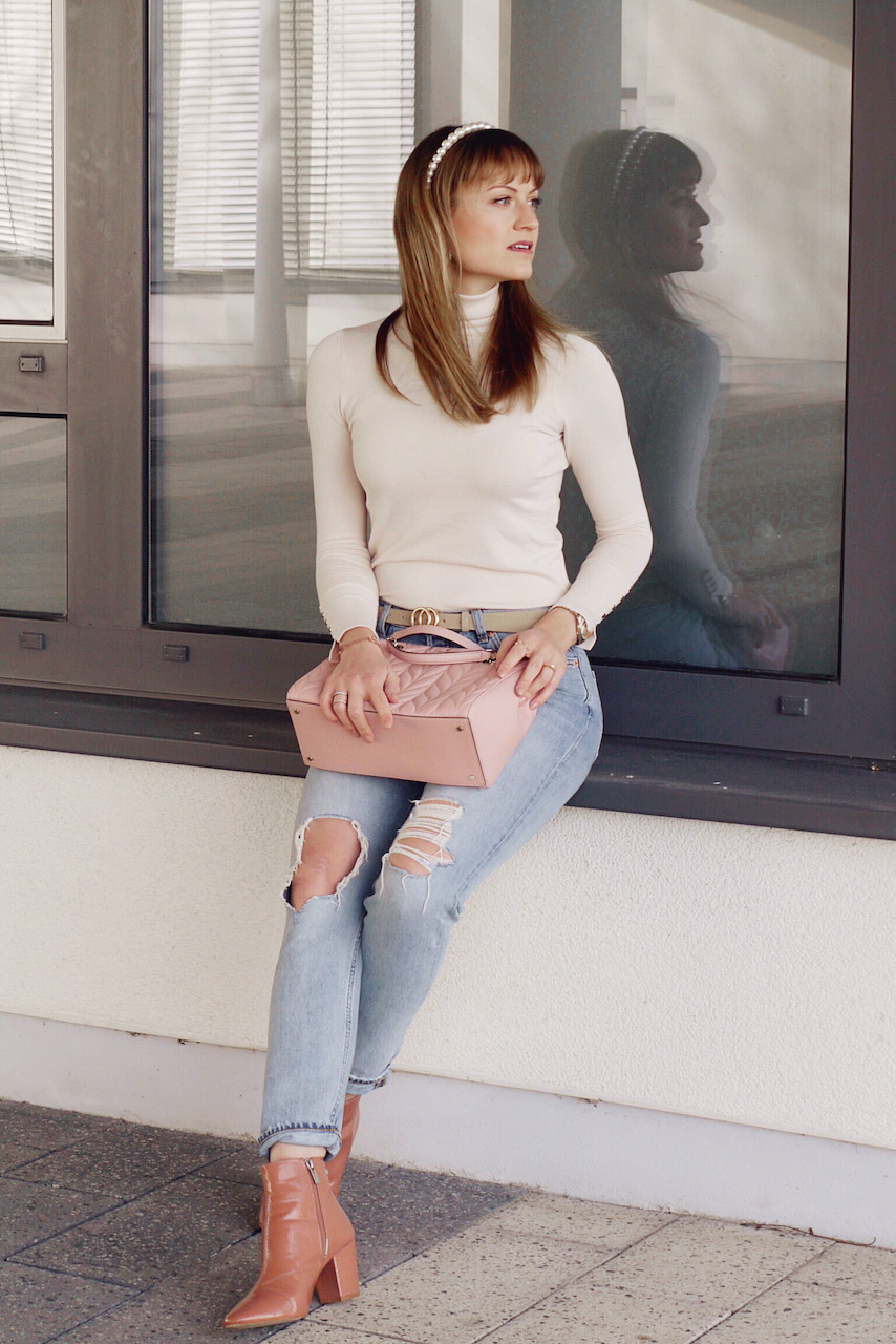 Girl in cream roll neck and distressed denim with pink boots and Kate Spade bag sitting in front of a window