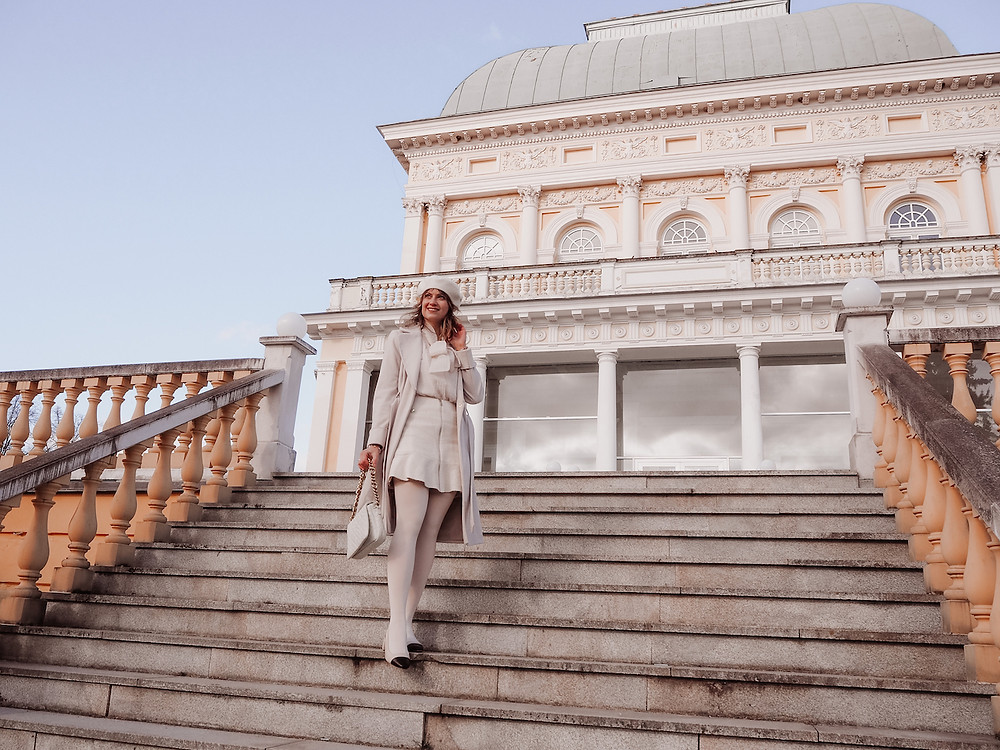 Young woman in an all-cream outfit consisting of a coat, a bouclé mini skirt, a pussy bow blouse, tights, a beret and slingback pumps in front of a magnificent building