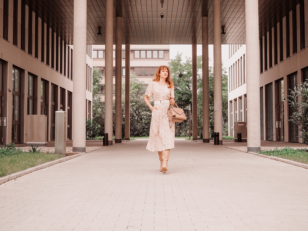 Young woman dressed in a floral midi dress, broad waist belt, beige boots and a brown bag in front of an office building